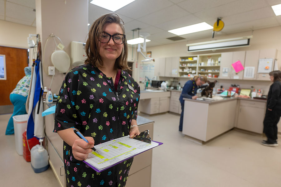 A community college student in scrubs smiles while holding a clipboard in a veterinary lab, representing the balance of working while in community college and pursuing a career change through hands-on education.