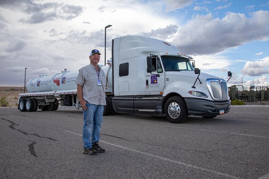 A commercial truck driver stands in front of a white semi-truck with a tanker trailer in an open lot under a partly cloudy sky. The image represents training for a Class A CDL, helping illustrate the types of CDL licenses and the difference between Class A and Class B CDL requirements in New Mexico.