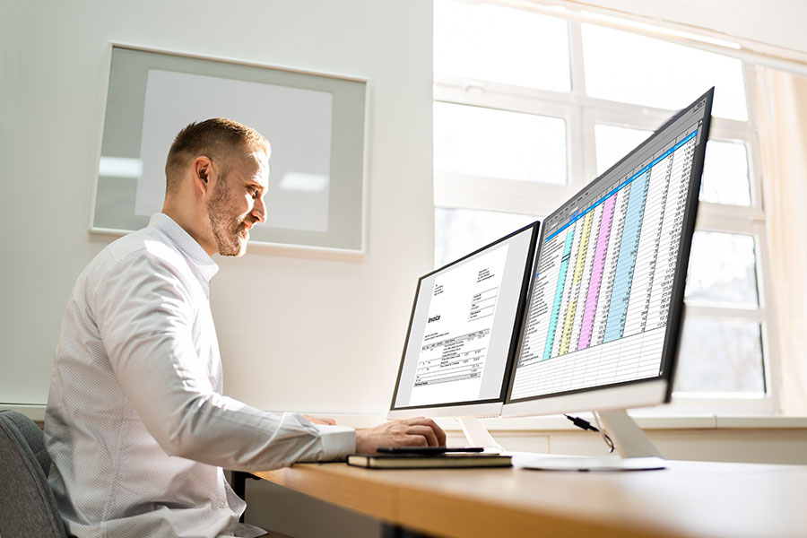A professional in an office reviews billing documents and colorful spreadsheets on dual monitors, representing the daily tasks of certified medical billing and coding specialists in a growing healthcare career field.